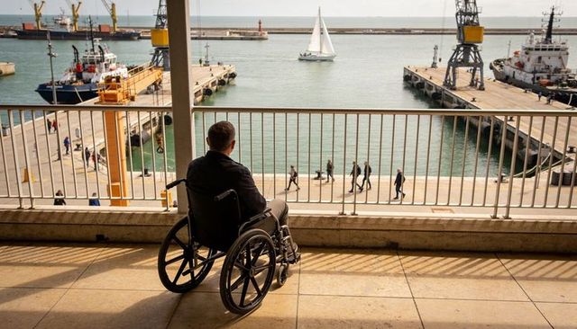 Contemplative man in wheelchair watching sailboat from sunlit harbor terrace overlooking dock cranes