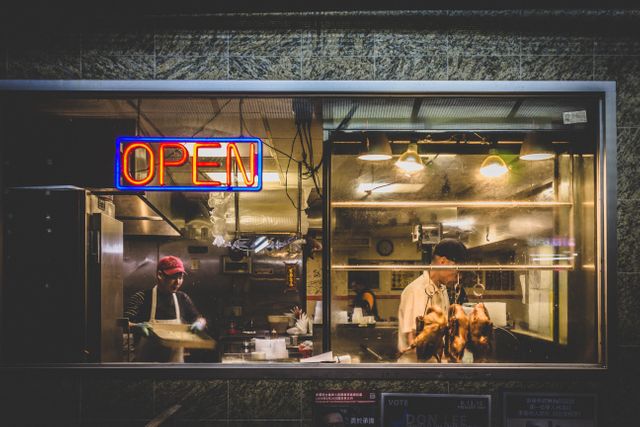 Two chefs preparing food in a restaurant kitchen during late hours. The neon 'Open' sign on the window indicates the restaurant is open to customers. Suitable for use in articles or promotions related to night dining, late-night eateries, culinary arts, and bustling urban nightlife.