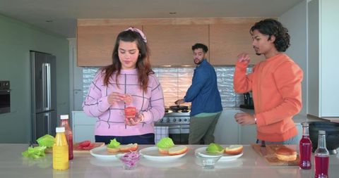 Friends preparing sandwiches at bright modern kitchen island, woman adding sauce
