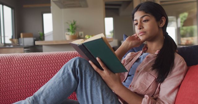 Image of biracial woman sitting on sofa and reading book. Relax, leisure and spending free time at home with technology concept.