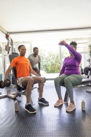 African american trainer coaching two clients lifting dumbbells on benches in bright gym