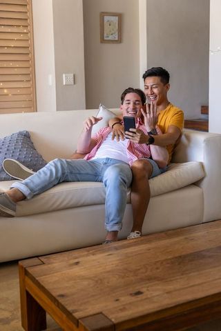Smiling interracial male couple sharing smartphone and relaxing on modern sofa