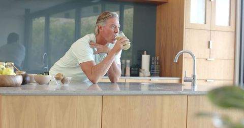 Mature man leaning on kitchen island drinking green smoothie in modern minimalist kitchen