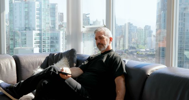 Adult man is relaxing on a comfortable couch in a modern city apartment during the daytime. He is casually dressed and holds a remote, appearing to be unwinding by watching TV or using another device. The large windows reveal a stunning urban cityscape, adding a sense of spaciousness and contemporary living. This image is perfect for themes of urban lifestyle, relaxation, home living, and modern apartments.