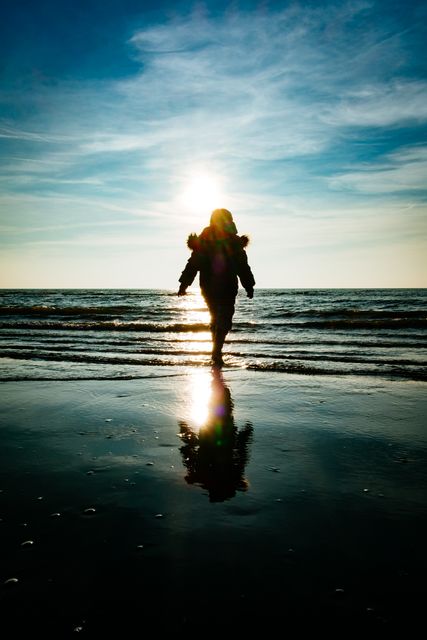 A person walking on the beach during sunset, casting a shadow and reflection on wet sand. Useful for themes of tranquility, nature, peaceful moments, solitude, introspection, and vacation destinations. Ideal for travel blogs, mental health articles, motivational content, and background images for websites or posters.