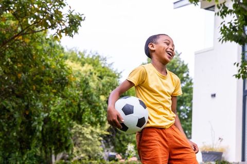 Laughing boy holding soccer ball in backyard with vibrant greenery and modern home