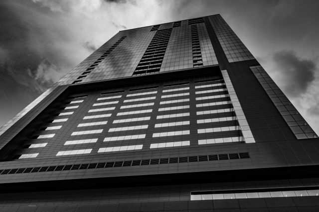 Captivating low angle view of a modern skyscraper under dramatic skies in black and white. Perfect for showcasing contemporary architecture, urban environments, business districts, and corporate advertisements. Useful for backgrounds, minimalist design concepts, and promotional materials related to construction and real estate.