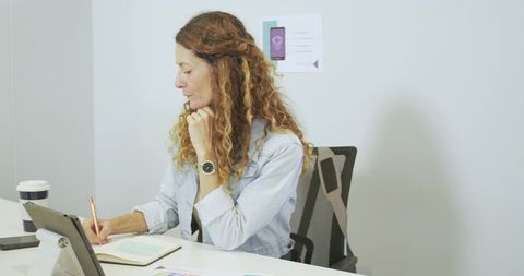 Mature woman taking notes at minimalist workspace with tablet coffee and documents