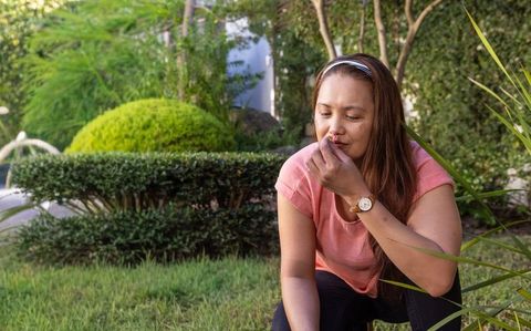 Asian woman kneeling on lawn smelling yellow flower in backyard garden, relaxed moment