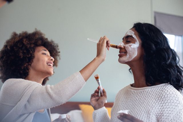 Happy biracial young woman with afro hair applying beauty cream on friend's face at home. Copy space, unaltered, friendship, togetherness, lifestyle, skin care and pampering concept.