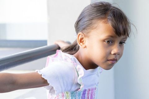 Young girl leaning on balcony railing, holding metal bar behind back, wearing pastel dress