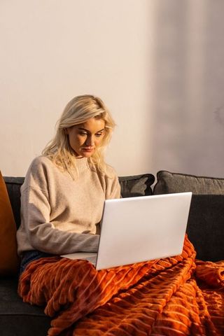 Woman working on laptop on couch wrapped in orange blanket during daylight