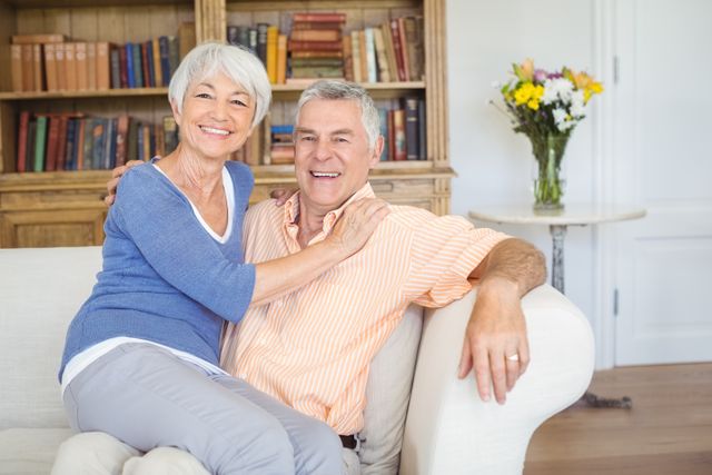 This image depicts a joyful senior couple relaxing in their living room. The woman is sitting on the man's lap, both smiling warmly. The background features a bookshelf filled with books and a table with a vase of flowers, adding to the cozy atmosphere. This image is ideal for use in advertisements, articles, or websites related to senior living, retirement, family life, and home comfort.