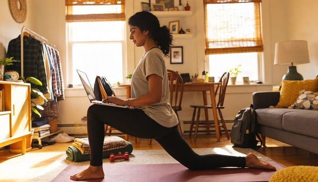 Woman lunging while typing on laptop in sunlit living room, multitasking remote work and workout