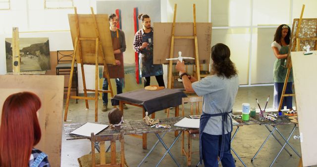 This depicts an arts class with students engaging in creative painting activities. The setting is an art studio with multiple easels and tables filled with painting supplies. It is ideal for use in content related to art education, creative workshops, learning environments, teamwork, young artists, and professional art studio setups.