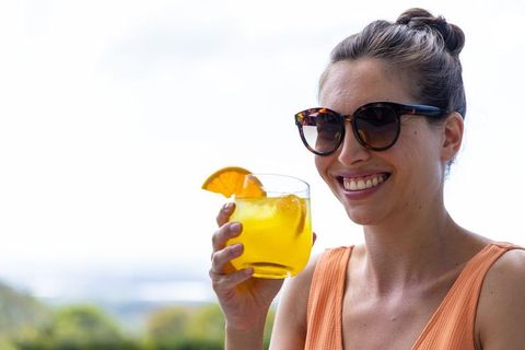 Smiling woman holding orange cocktail on sunny terrace wearing tortoiseshell sunglasses