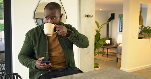 African American Man Sipping Coffee at Kitchen Island While Checking Phone, Morning
