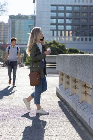 Young woman walking on sunny urban bridge talking on smartphone holding coffee