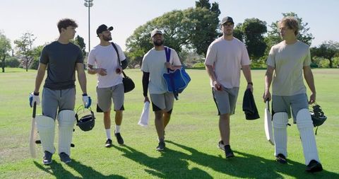Diverse male cricket team walking across sunlit field carrying bats, pads and helmets in sportswear