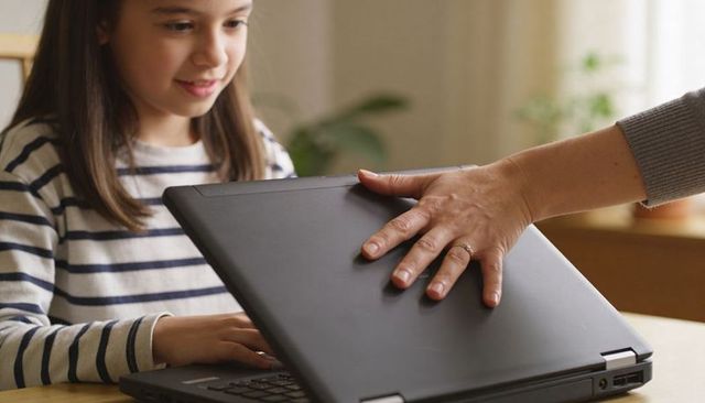 Child typing on laptop as caregiver closing lid with ringed hand parental control moment