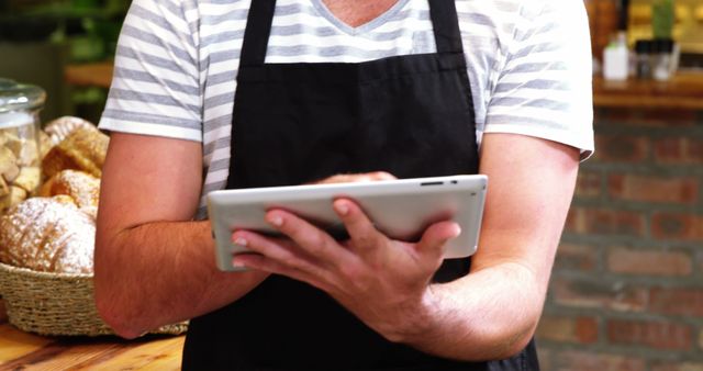 Bakery worker stands in shop holding a tablet, likely managing inventory or taking orders. Background includes bread and pastries. Suitable for illustrating modern technology use in small businesses, retail environment management, and bakery promotional materials.