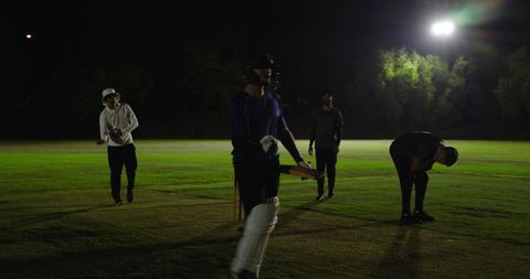 Night cricket practice under floodlights: batter walking in helmet and pads with teammates
