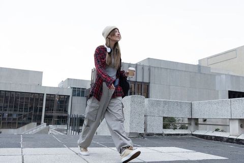Young woman walking rooftop terrace holding coffee cup wearing beanie and headphones