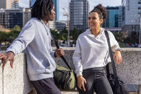 Sunlit diverse friends leaning on urban railing holding duffel bag and smartphone