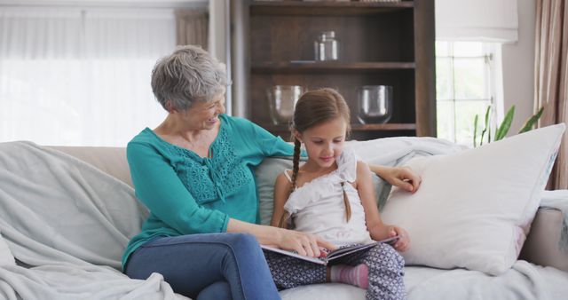 Happy caucasian grandmother and granddaughter sitting on sofa, reading book together. Lifestyle, domestic life, family, and togetherness.