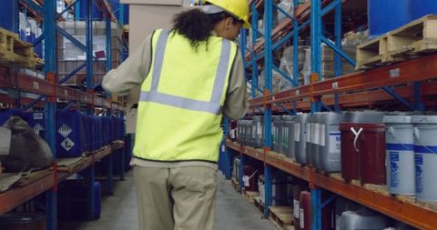 Warehouse worker walking aisle wearing hi-vis vest and hard hat near labeled drums