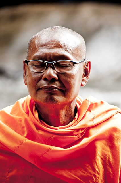 Buddhist monk in an orange robe meditating with eyes closed. Wearing glasses, he exudes a sense of peace and tranquility. Useful for topics related to spirituality, meditation, mindfulness, and inner peace.