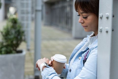 Asian woman checking smartwatch while holding takeaway coffee on urban walkway