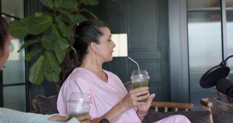 Woman speaking on casual podcast holding green smoothie on sofa with microphone