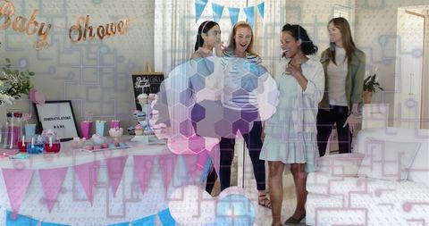 Friends celebrating baby shower while mother-to-be holding baby outfit with pastel balloons and dess