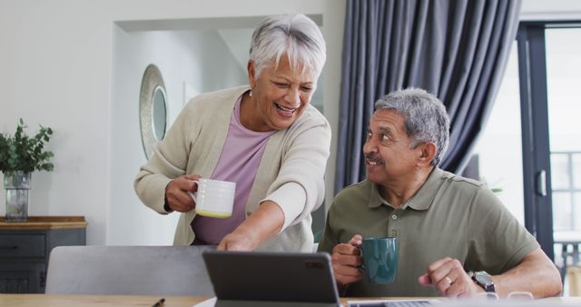 Happy senior biracial couple talking with coffee and using laptop. Spending quality time at home, retirement and lifestyle concept.