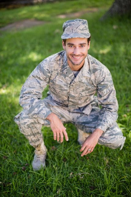 Young soldier in camouflage uniform smiling while squatting in a park. Ideal for use in articles about military life, recruitment campaigns, or veteran stories. Can also be used in promotional materials for military events or community support programs.