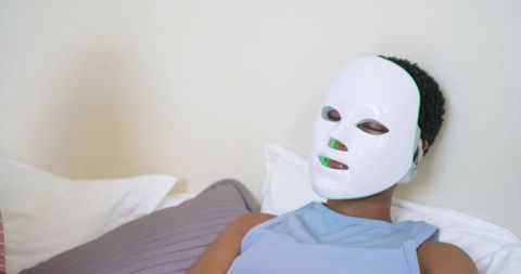 African american woman relaxing with led light therapy facial mask on bed, home skincare wellness
