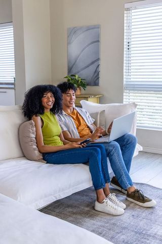 African american woman and asian man on sofa using laptop and remote in bright modern living room