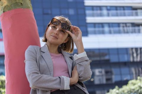 Young businesswoman leaning on pink column adjusting sunglasses near glass office facade