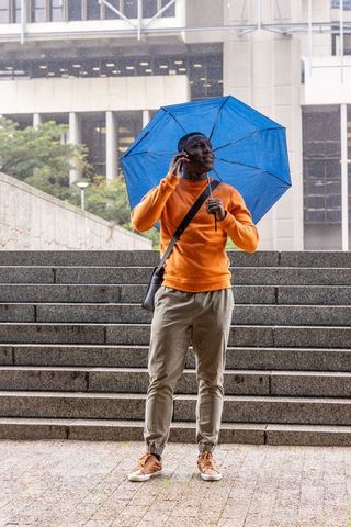 African american commuter standing in rain holding blue umbrella and talking on phone