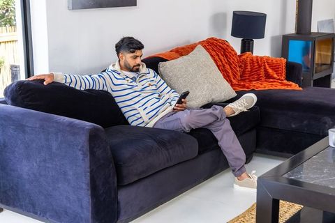 Indian man relaxing on navy sectional sofa checking smartphone in modern living room