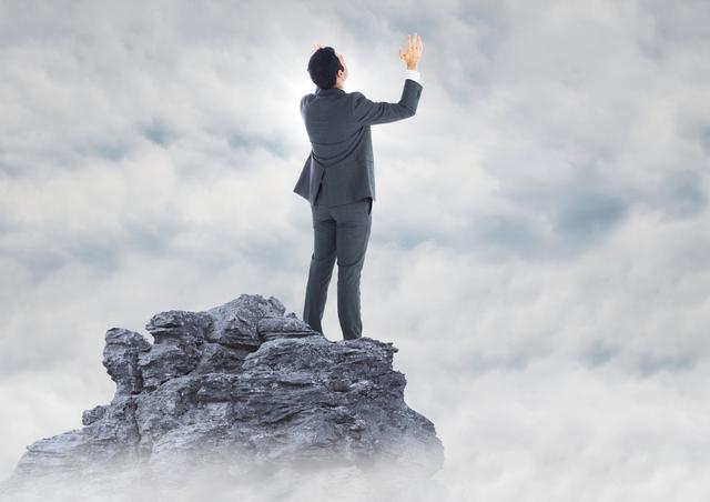 Businessman in a suit standing on a rocky mountain peak with hands raised in triumph, surrounded by clouds and blue sky. Ideal for concepts of success, achievement, reaching goals, professional milestones, corporate motivation, and inspirational business stories.