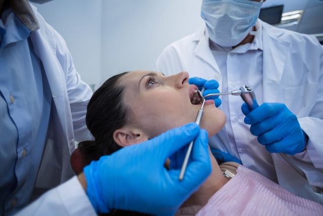Dentists are examining a female patient using dental tools in a clinical setting. The patient is lying back in a dental chair while the dentists, wearing blue gloves and white coats, perform a dental check-up. This image can be used for promoting dental health services, illustrating dental procedures, or in educational materials about oral hygiene and healthcare.