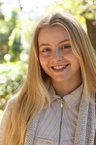 Young blonde woman smiling in sunlit park wearing striped collared zipped shirt