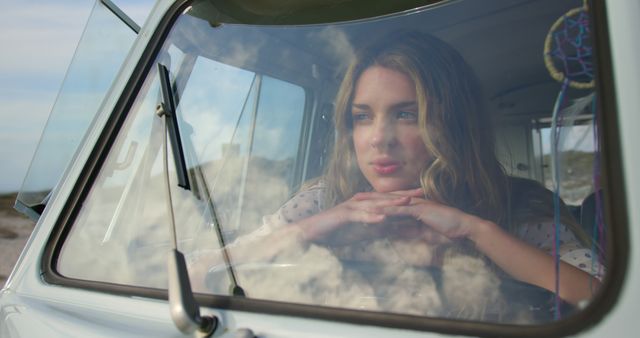 A woman with hands under chin is gazing dreamily through a windshield of a vintage car, with clouds reflecting on the glass. Ideal for travel blogs, lifestyle articles, and advertisements conveying themes of daydreaming, relaxation, or nostalgia.