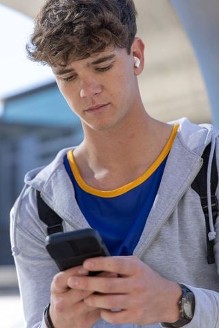 Teen student checking smartphone while wearing earbuds and backpack near campus