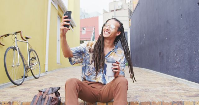 Front view of a biracial man with long dreadlocks out and about in the city on a sunny day, sitting in the street, drinking coffee, taking a selfie with his smartphone, with his bicycle leaning against the wall next to him in slow motion.