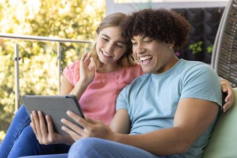 Happy diverse couple video-calling and laughing on sunlit balcony sofa holding tablet