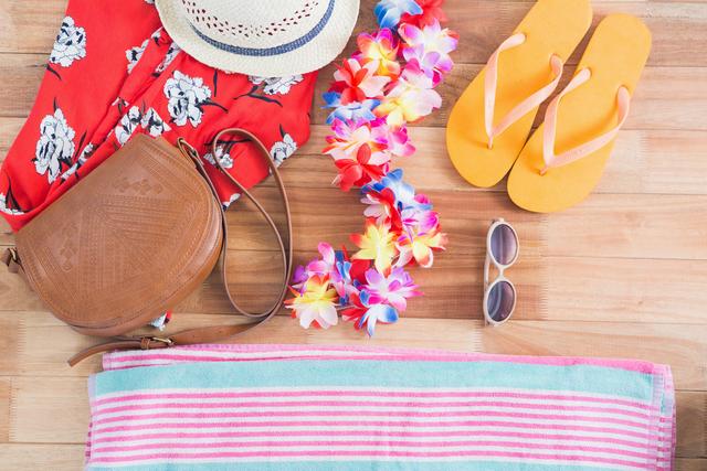 Flat lay of tropical beach holiday accessories on wooden floor. Includes a straw hat, floral lei, orange flip-flops, sunglasses, brown leather bag, striped towel, and red floral dress. Perfect for travel blogs, vacation planning websites, summer fashion promotions, and tropical getaway advertisements.