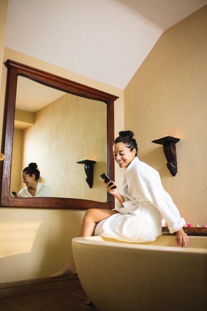 This image depicts a smiling biracial young woman wearing a bathrobe and using a smartphone while sitting on a bathtub in a spa bathroom. The setting suggests a relaxing and luxurious environment, ideal for promoting wellness, self-care, and modern lifestyle themes. Perfect for use in advertisements for spas, wellness retreats, beauty products, or technology in lifestyle contexts.
